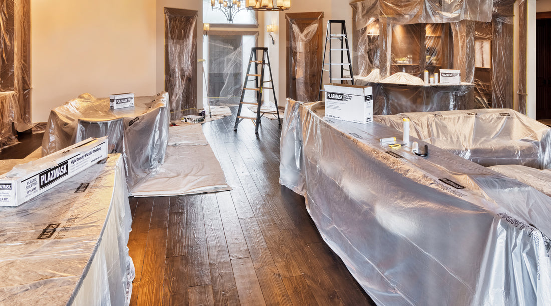 A wide shot of a living room with all furniture and doorways covered in masking film to protect from paint and debris during renovations. Two ladders can be seen in the background. 2 White boxes of Plazmask plastic sheeting and 2 white boxes of Plazmask masking film can be seen scattered through the room.