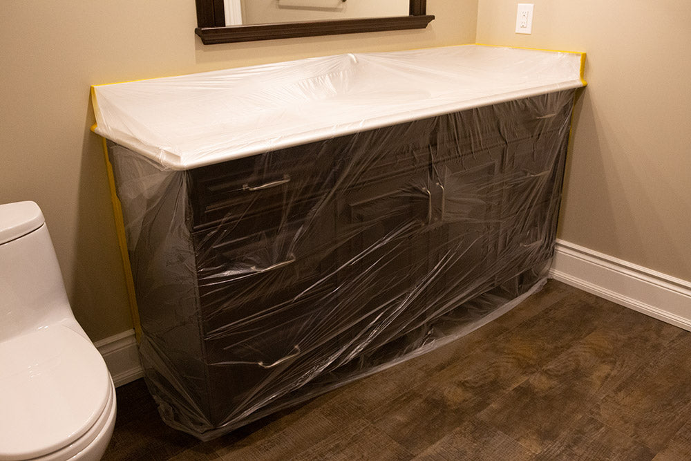 A black bathroom cabinet with a white countertop that is mounted to a wall below a mirror is covered in Plazmask masking film.
