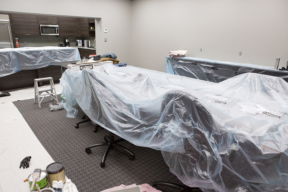 Office chairs and a boardroom table can be seen covered in masking film, a cabinet in the back right is covered in masking flim as well, a kitchen counter in the back left is also protected with masking film. Paint cans and brushes are in the left foreground, with a small step ladder sitting in between the paint cans and kitchen counter. 