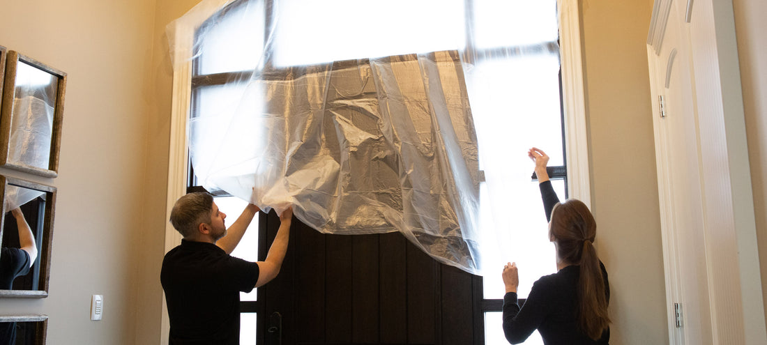 A man in a black shirt and a woman in a black shirt are applying masking film to a black front door and windows,
